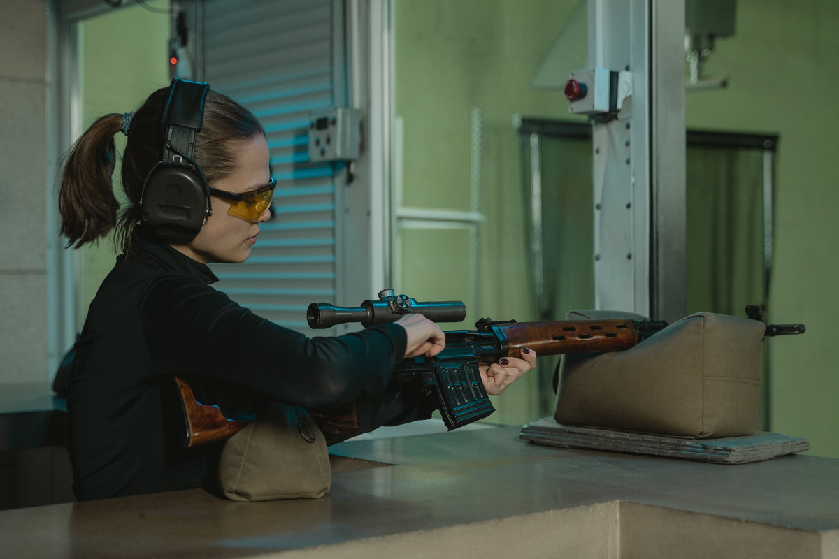 Person wearing hearing and eye protection aims a scoped rifle at an indoor shooting range.
