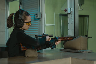 Person wearing hearing and eye protection aims a scoped rifle at an indoor shooting range.